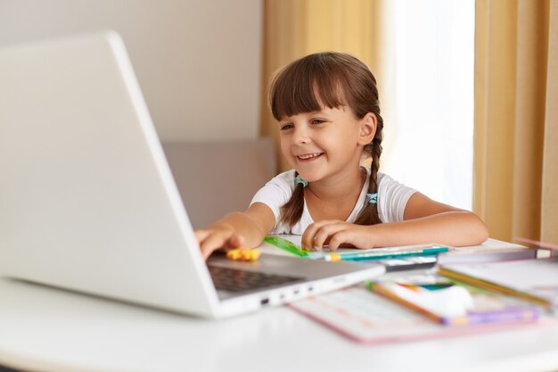Kryty strzał szczęśliwy pozytywny ciemnowłosy uczeń pozujący w domu, patrzący na komputer przenośny z uroczym uśmiechem, kształcenie na odległość, lekcję online.