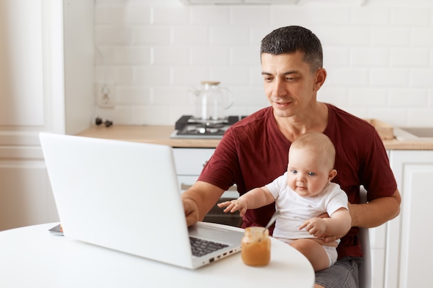 Kryty strzał uśmiechający się przystojny mężczyzna freelancer na sobie bordową koszulkę, pozowanie w białej kuchni, siedząc przed laptopem z dzieckiem w rękach, pisząc na notebooku.