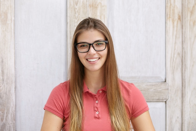 Bezpłatne zdjęcie kryty strzał z piękną młodą kobietą rasy kaukaskiej na sobie koszulkę polo i prostokątne okulary uśmiechając się radośnie podczas pozowanie na białym tle