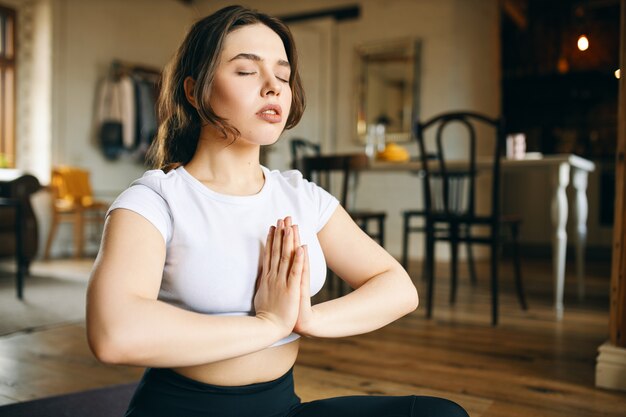 Kryty ujęcie spokojnej młodej kobiety siedzącej w domu w pozycji medytacyjnej z zamkniętymi oczami, wykonującej gest namaste, medytującej, skupiającej umysł na oddychaniu, trenującej uwagę i świadomość