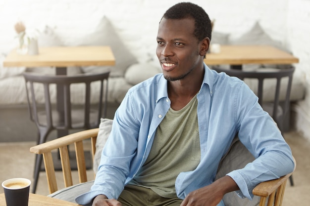 Bezpłatne zdjęcie kryty ujęcie uśmiechniętego czarnego nieogolonego mężczyzny w koszuli, siedzącego przy drewnianym krześle w kawiarni, pijącego gorącą kawę lub cappuccino, patrzącego na kogoś z uśmiechem i przyjemnej rozmowy z przyjacielem