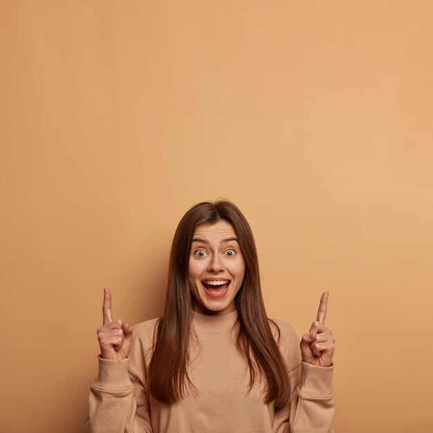 Kryty ujęcie zadowolonej, zachwyconej kobiety wskazuje powyżej palcami wskazującymi, radośnie się śmieje, reklamuje chłodną pustą przestrzeń, nosi luźny sweter