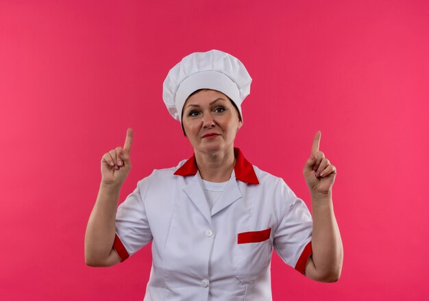 kucharz w średnim wieku w mundurze szefa kuchni wskazuje palcami w górę na odosobnionej różowej ścianie