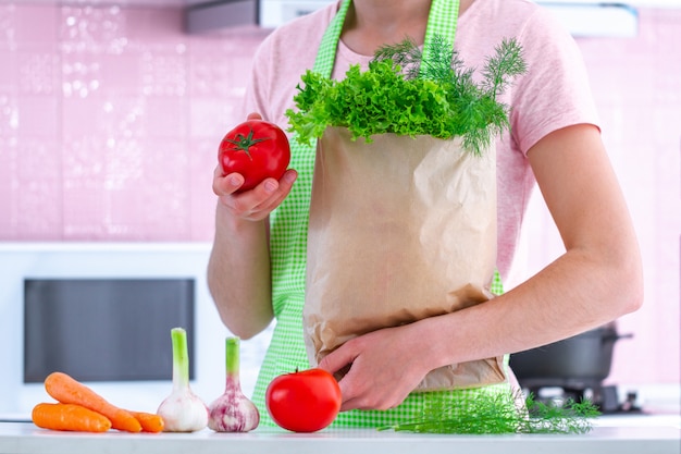Kulinarna kobieta w fartuchu trzyma rzemieślniczą papierową torbę pełno świezi organicznie warzywa przy kuchnią. zdrowe jedzenie i zbilansowana dieta, czyste jedzenie