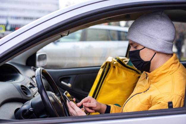 Kurier W Samochodzie Z Czarną Maską Medyczną Ma Na Telefonie, Plecak Z Dostawą Na Siedzeniu. Usługa Dostawy Jedzenia