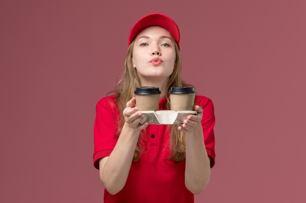 kurierka w czerwonym mundurze trzyma brązowe kubki do kawy wysyłające pocałunki na różowym, mundurze pracownika służby pracy