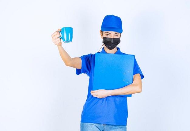 Kurierka w niebieskim mundurze i czarnej masce na twarz trzymająca niebieską teczkę i dzieląca się filiżanką herbaty z kolegą.