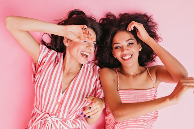 Kuzyni w cudownym nastroju leżą na podłodze w świetnym nastroju. Dziewczyny pozują ze szczerymi uśmiechami w różowych ubraniach do portretu z bliska.