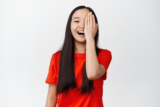 Ładna azjatycka kobieta zakrywa jedną stronę twarzy i uśmiecha się śmiejąc się beztrosko stojąc na białym tle w czerwonej koszulce
