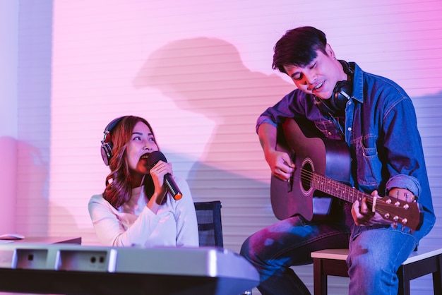 Ładna azjatycka piosenkarka nagrywająca piosenki za pomocą mikrofonu studyjnego z mężczyzną grającym na gitarze w niebieskim i czerwonym świetle. występ i show w branży muzycznej. sesja w duecie.