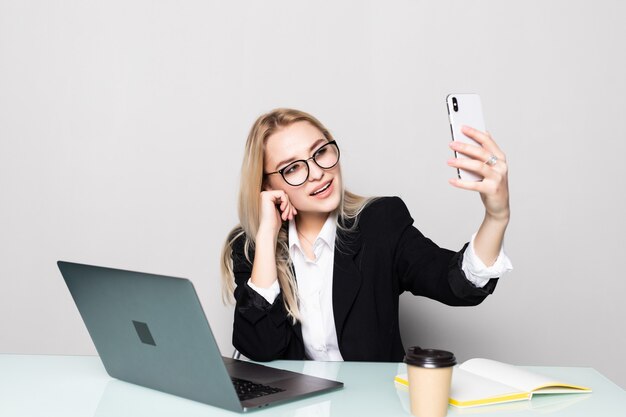 Ładna biznesowa kobieta trzyma jej telefon komórkowego z jedną ręką i robi rozmowie wideo przy biurowym biurkiem w biurze