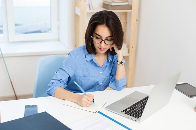 Ładna brunetka dziewczyna w niebieskiej koszuli pracuje przy stole w biurze. Pisze poważnie w zeszycie.
