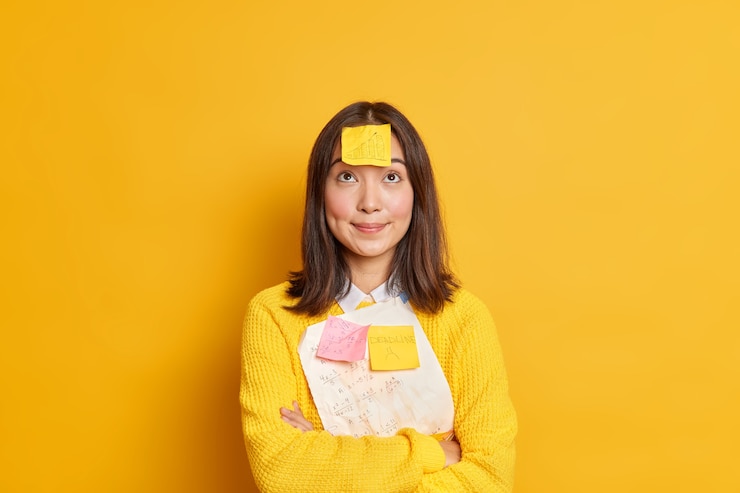 Bezpłatny plik Zdjęcie ładna brunetka, młoda azjatycka studentka próbuje zapamiętać materiał do egzaminu, ma naklejkę z informacją o terminie przyklejoną do siebie, aby skoncentrować się nad stojakami skrzyżowanymi rękami