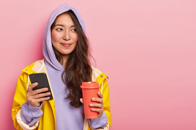 Ładna brunetka młoda kobieta o naturalnym pięknie, używa nowoczesnego telefonu komórkowego do rozmów z przyjaciółmi, trzyma kawę na wynos