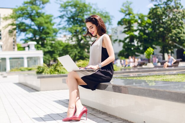 Ładna brunetka w szaro-czarnej sukience i winnych obcasach siedzi w parku w mieście. Pisze na laptopie i wygląda na zadowoloną z pracy na zewnątrz.