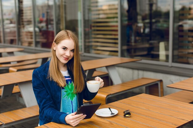 Ładna dziewczyna siedzi przy stole na tarasie. Trzyma filiżankę kawy i tabletkę. Ona uśmiecha się do kamery.