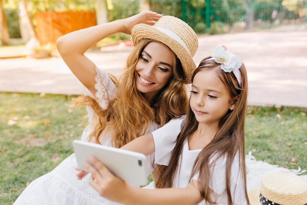 Ładna dziewczyna w białej sukni, trzymając smartfon i robiąc selfie z roześmianą mamą idąc ulicą. Zewnątrz portret zadowolony młoda kobieta w kapeluszu pozowanie, podczas gdy brunetka córka robi zdjęcie.