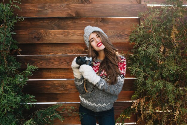 Ładna dziewczyna z długimi włosami i śnieżnobiałym uśmiechem w czapka i rękawiczki na drewnianym zewnątrz. Nosi sweter, trzyma aparat, uśmiecha się.