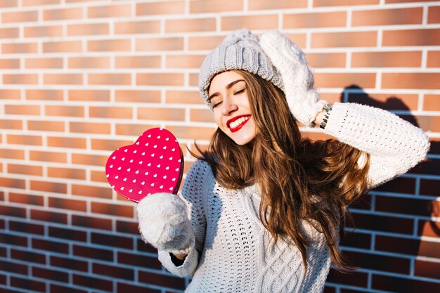 Ładna dziewczyna z długimi włosami w ciepły sweter i czapka na ścianie na zewnątrz. W rękawiczkach trzyma czerwone serce, z zamkniętymi oczami wygląda na zadowoloną.