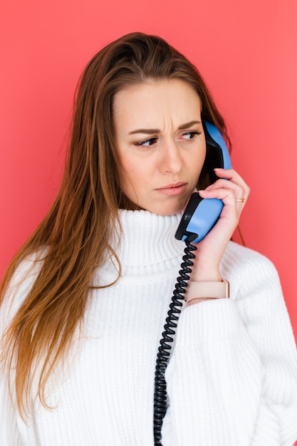 Bezpłatne zdjęcie Ładna europejska kobieta w swobodnym białym swetrze trzyma telefon stacjonarny po rozmowie z ciekawską zdezorientowaną poważną twarzą