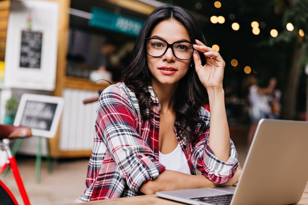 Ładna kobieta freelancerka nosi modne okulary pozujące w mieście blur. Elegancka czarnowłosa dziewczyna za pomocą laptopa w dobry dzień.