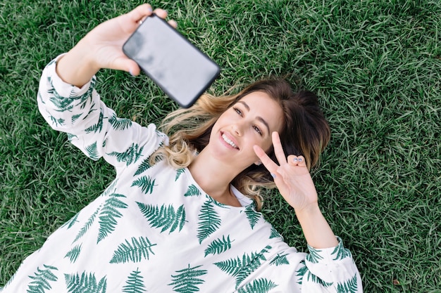 Ładna kobieta, kładąc na trawie w parku i robiąc selfie na swoim telefonie i pokazuje znak pokoju