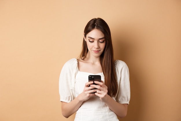Ładna kobieta wysyła SMS-y na ekranie odczytu telefonu komórkowego smartfona i uśmiecha się stojąc na beżowym tle