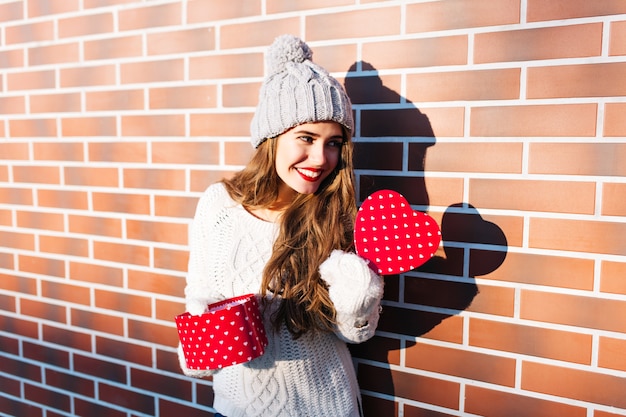 Ładna młoda dziewczyna w czapkę, ciepły sweter i rękawiczki na ścianie na zewnątrz. Trzyma w dłoniach otwarte pudełko z sercem, uśmiechając się na bok.