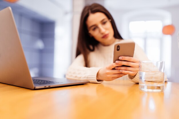 Ładna młoda kobieta siedzi w kuchni z laptopem ma wideo połączenie na jej telefonie