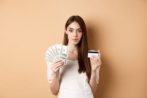 Ładna młoda kobieta w białej bluzce, pokazująca banknoty dolarowe i płatność zbliżeniową plastikową kartą kredytową...