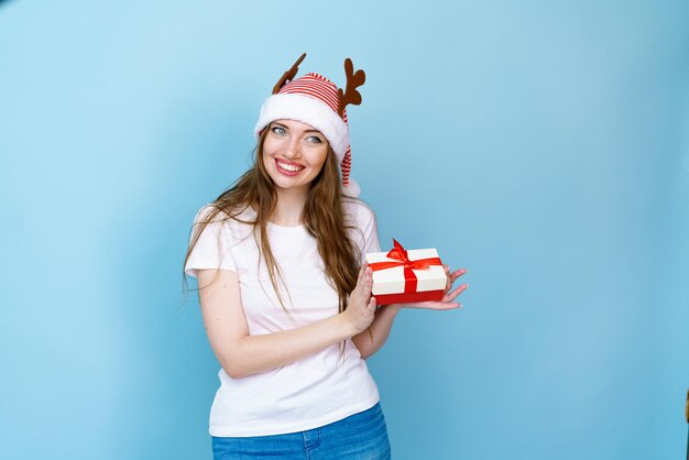 Ładna młoda kobieta w bożonarodzeniowe poroże jelenia i białą koszulkę na białym tle na niebieskim tle trzymając ...
