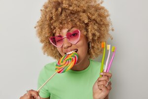 Ładna nastolatka gryzie słodycze wielokolorowe karmelowe cukierki trzyma szczoteczki do zębów nie może odmówić jedzenia szkodliwego jedzenia dla zębów nosi różowe odcienie serca i niezobowiązującą zieloną koszulkę pozuje w pomieszczeniach