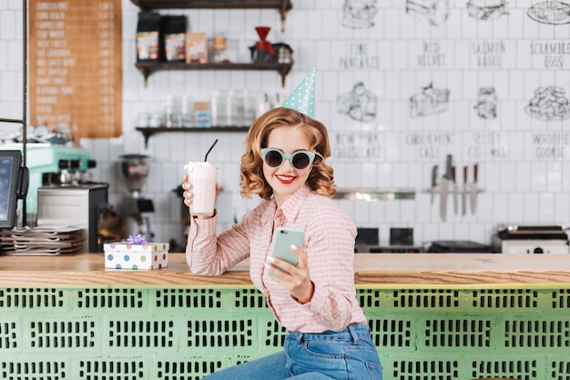 Ładna pani w okularach przeciwsłonecznych i czapce urodzinowej siedząca przy barze z koktajlem mlecznym i telefonem komórkowym w rękach, podczas gdy szczęśliwie spędza czas w kawiarni