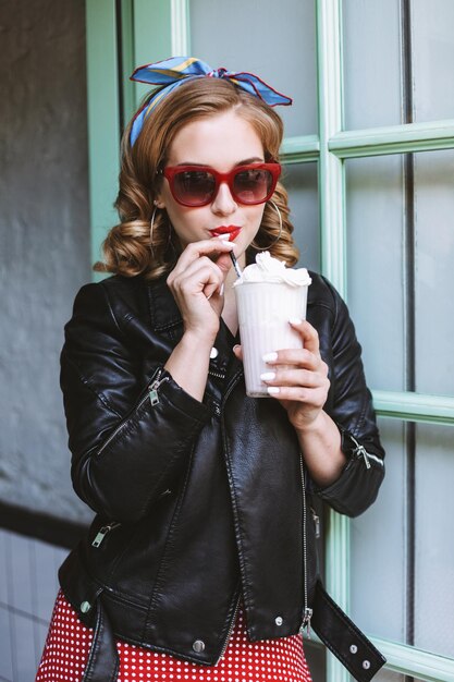 Ładna pani w okularach przeciwsłonecznych i skórzanej kurtce stojąca w pobliżu drzwi w kawiarni i pijąca koktajl mleczny