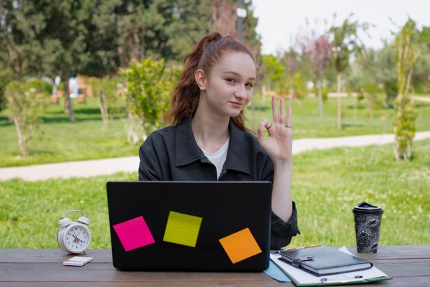 Ładna studencka freelancerka pracująca przy laptopie na zewnątrz, pokazująca, że wszystko jest w porządku