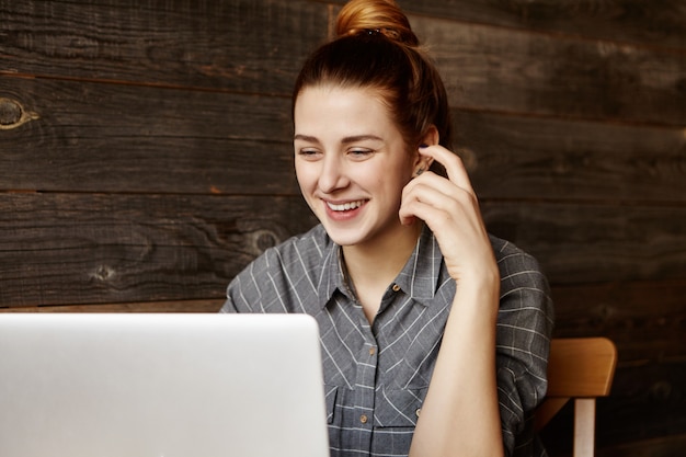 Bezpłatne zdjęcie Ładna studentka z uroczym uśmiechem siedząca przed laptopem, prowadząca rozmowę wideo z przyjacielem, śmiejąca się radośnie, korzystając z bezprzewodowego szybkiego łącza internetowego w kawiarni podczas przerwy na kawę