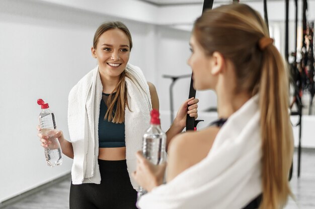 Ładne młode dziewczyny fitness trzymają butelki z wodą, patrzą na siebie i uśmiechają się