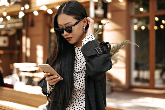 Ładnie opalona kobieta w czarnym trenczu i białej sukience pozuje na zewnątrz i wysyła wiadomości do telefonu Portret pięknej azjatyckiej damy w okularach przeciwsłonecznych