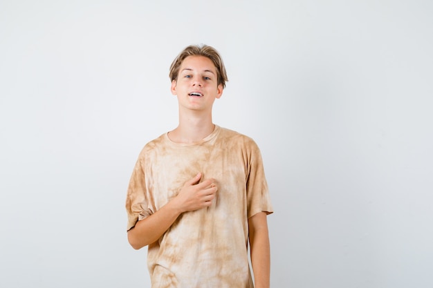 Ładny nastolatek chłopiec trzymając rękę na klatce piersiowej w t-shirt i patrząc dumny, widok z przodu.