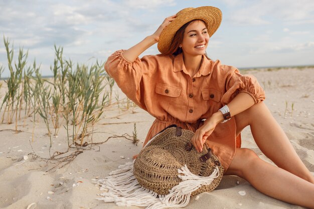 Ładny uśmiechający się dziewczyna w stylowej lnianej sukience relaksujący na wieczornej plaży na białym piasku. Kapelusz słomkowy, torebka boho. Święta mod.