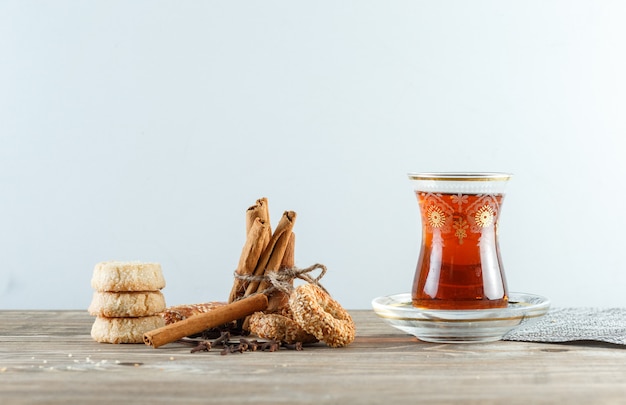 Laski cynamonu z herbatnikami, goździkami, szklanką herbaty, podkładką widok z boku na drewnianej i białej ścianie