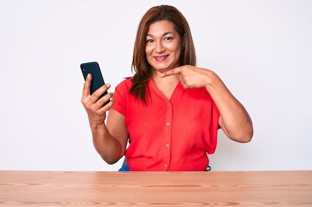 Latynoska brunetka w średnim wieku, używająca smartfona, siedząca na stole, uśmiechnięta, szczęśliwa, wskazująca ręką i palcem