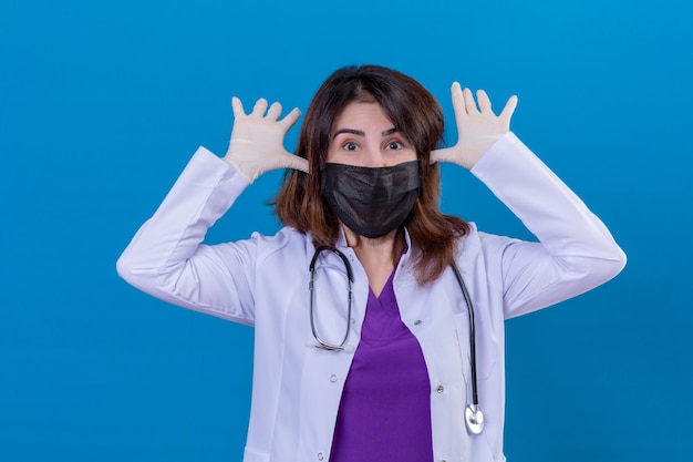Bezpłatne zdjęcie lekarka w średnim wieku ubrana w biały fartuch w czarnej ochronnej masce na twarz i ze stetoskopem uśmiechnięta wesoło grająca w okiem z opuszczonymi rękami i szczęśliwa stojąca na niebieskim tle