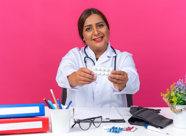 Bezpłatne zdjęcie lekarka w średnim wieku w białym fartuchu ze stetoskopem trzymająca blister z pigułkami patrząca uśmiechnięta radośnie siedząca przy stole z folderami biurowymi na różowym tle