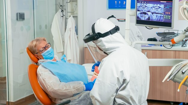 Lekarz dentysta w kombinezonie pokazującym prawidłową higienę jamy ustnej z wykorzystaniem makiety szkieletu zębów podczas pandemii koronawirusa. Zespół medyczny ubrany w kombinezon ochronny, maskę na twarz i rękawiczki.