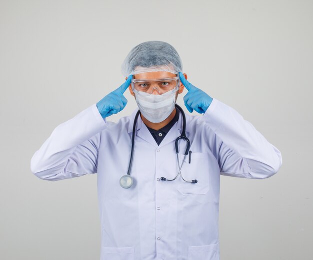 Lekarz dotyka skroni palcami w białym fartuchu, masce, kapeluszu i patrzy uważnie