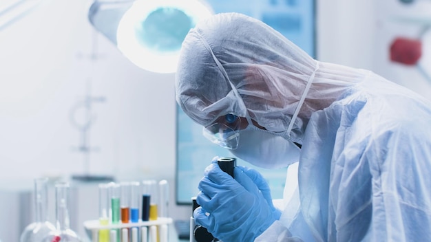 Lekarz naukowiec ze sprzętem PPE patrzący na mikroskop medyczny analizujący próbkę wirusa podczas eksperymentu opracowywania szczepionki w szpitalnym laboratorium biochemicznym. Koncepcja medycyny