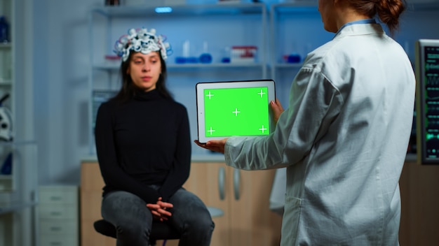 Bezpłatne zdjęcie lekarz neurolog trzymając tablet z zielonym ekranem w laboratorium badań neurologicznych. naukowiec korzystający z makiety wyświetlacza klucza chromatycznego na białym tle pulpitu, podczas gdy pacjent z zestawem słuchawkowym eeg macha w tle