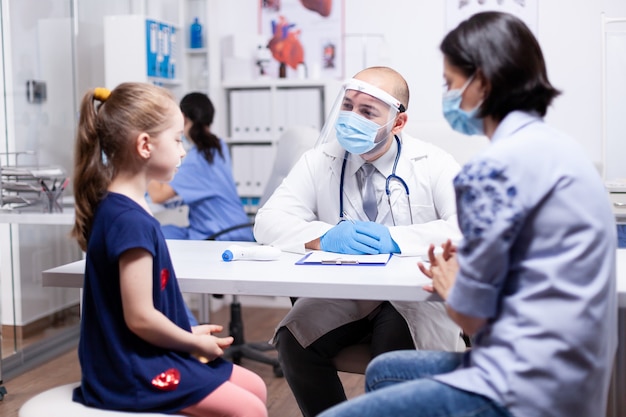 Lekarz nosi maskę podczas globalnej pandemii rozmawiając z dzieckiem podczas konsultacji. Specjalista zdrowia pediatra udzielający porad w zakresie opieki zdrowotnej leczenie w sprzęcie ochronnym.