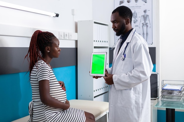 Lekarz pokazuje tablet z pionowym zielonym ekranem do kobiety w ciąży. pacjent spodziewa się dziecka patrząc na klucz chrominancji z izolowanym szablonem i tłem makiety dla opieki zdrowotnej.